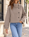 Veste courte femme beige à col montant asymétrique, fermeture boutonnée noire, poches latérales et manches longues avec pattes boutonnées